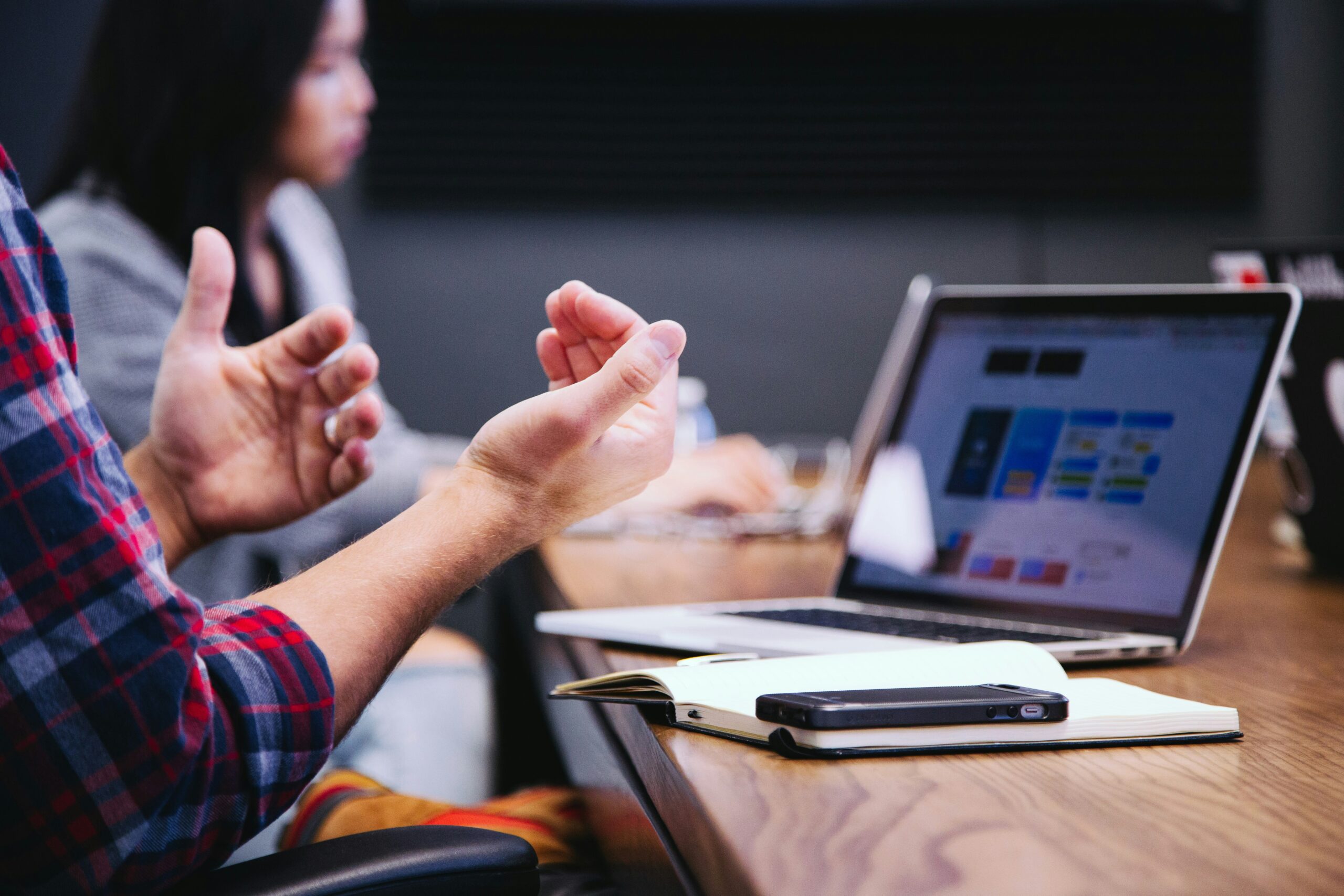 A man sitting in front of a laptop, gesturing with his hand as if explaining something.